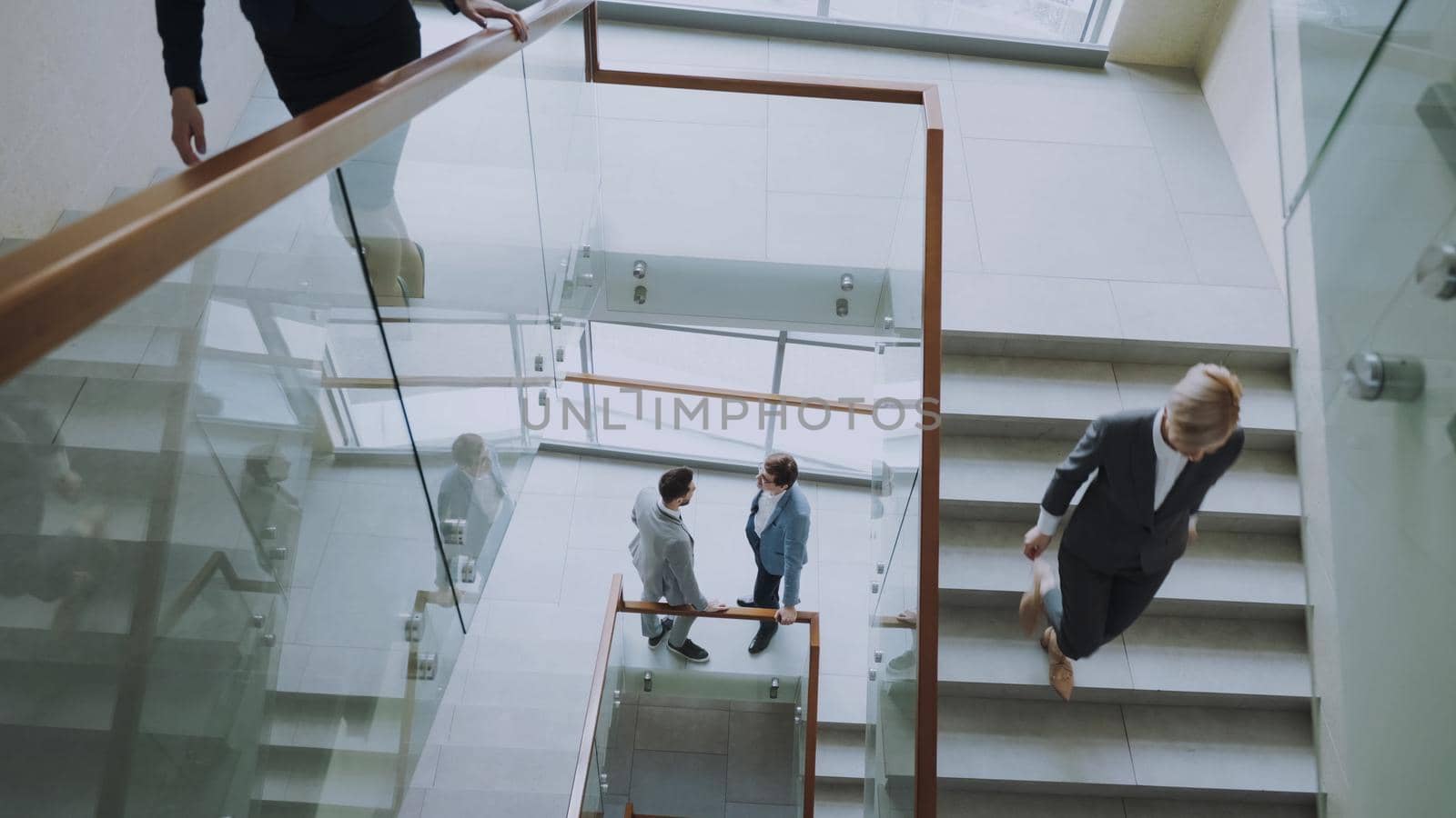 Top view of two businessmen meet at staircase in modern office center and talking while female colleagues walking stairs by silverkblack