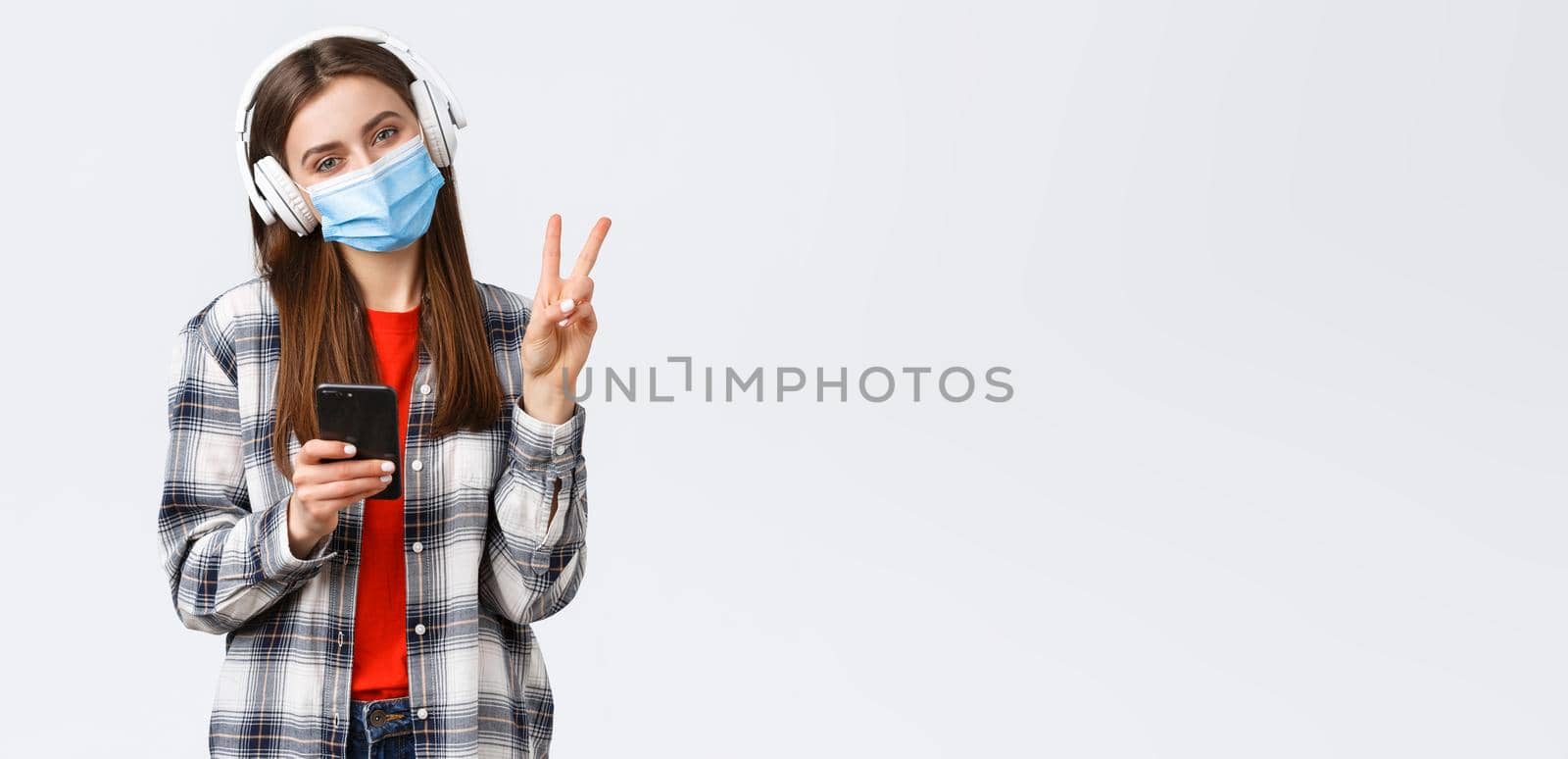 Social distancing, leisure and lifestyle on covid-19 outbreak, coronavirus concept. Cute silly teenage girl in medical mask and headphones, show peace sign, relaxing with music, hold mobile phone.