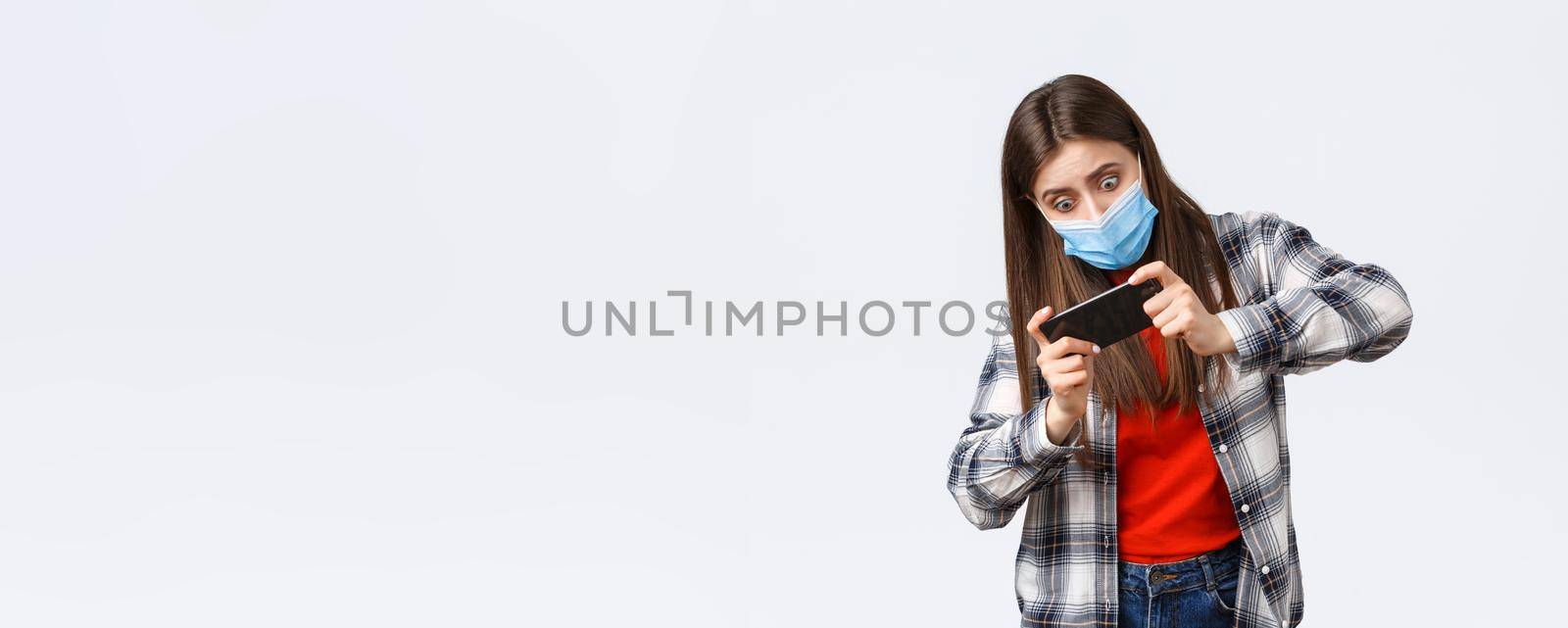 Different emotions, covid-19 pandemic, coronavirus self-quarantine and social distancing concept. Focused and entertained woman in medical mask playing mobile game, tilting body, difficult level by Benzoix