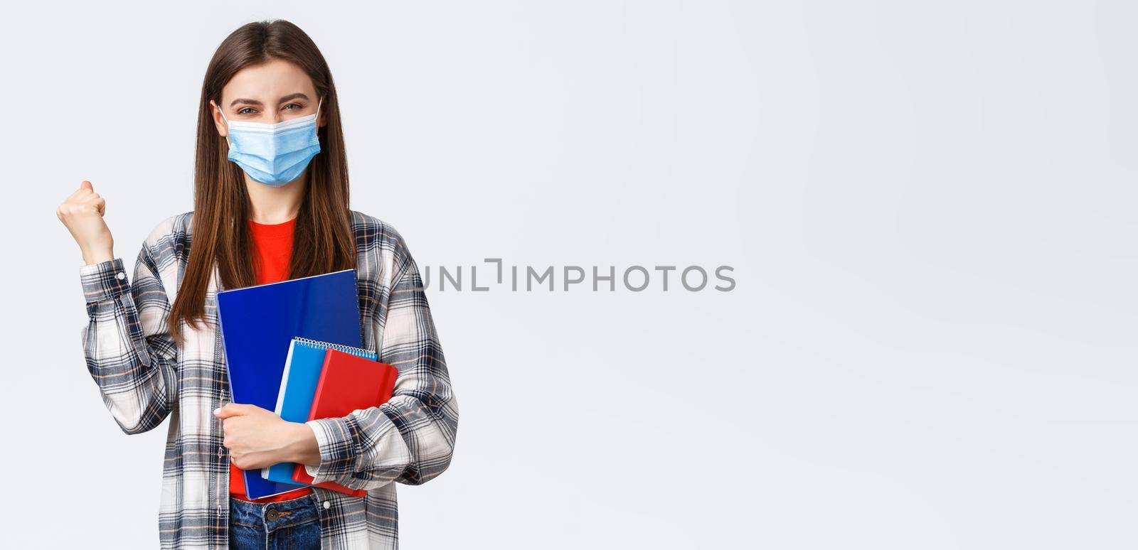 Coronavirus pandemic, covid-19 education, and back to school concept. Rejoicing, cheerful female student in medical mask happy entering cool university, fist pump in success, holding notebooks.