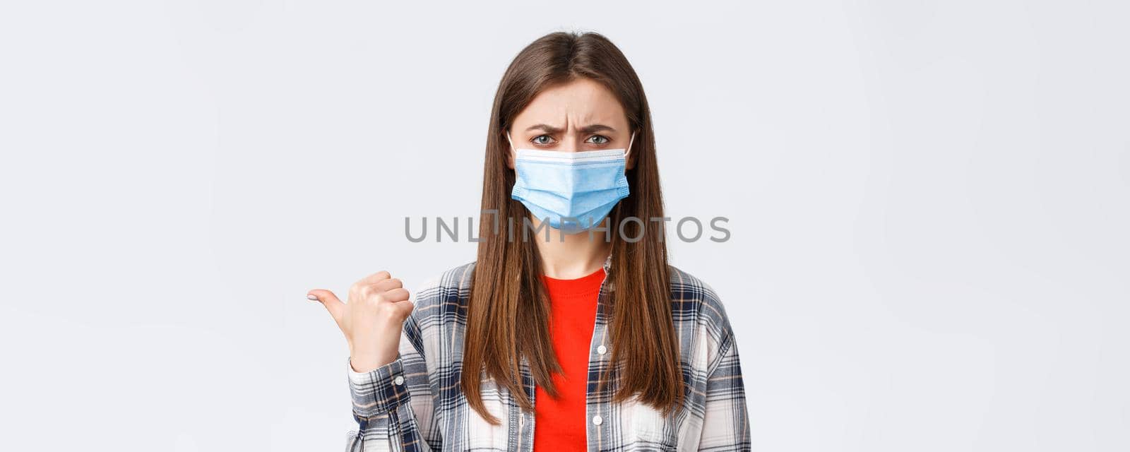 Coronavirus outbreak, leisure on quarantine, social distancing and emotions concept. Upset and disappointed young woman in medical mask frowning, condemn smth, pointing finger left displeased.