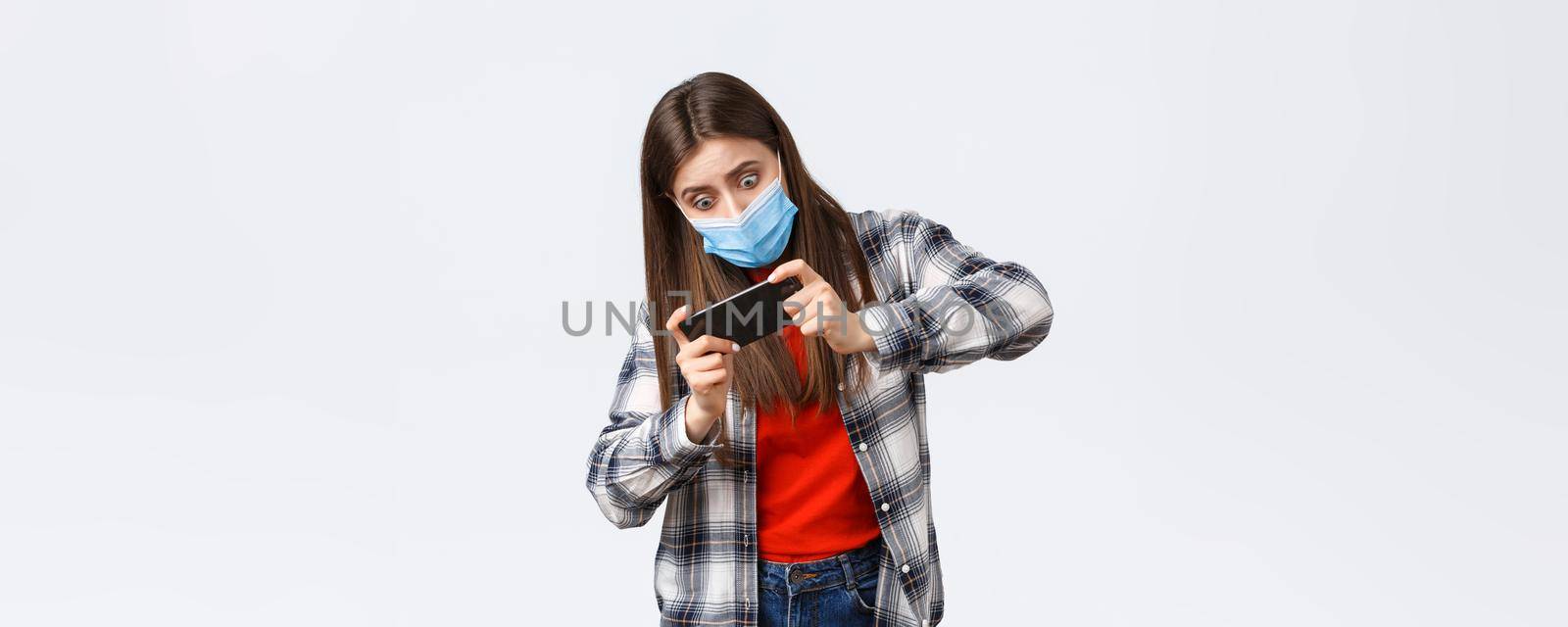 Different emotions, covid-19 pandemic, coronavirus self-quarantine and social distancing concept. Focused and entertained woman in medical mask playing mobile game, tilting body, difficult level.