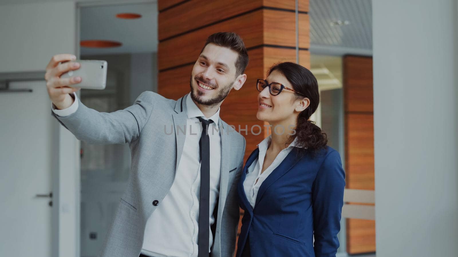 Happy businessman and his female colleague taking a selfie on smarphone camera for social media in modern office by silverkblack
