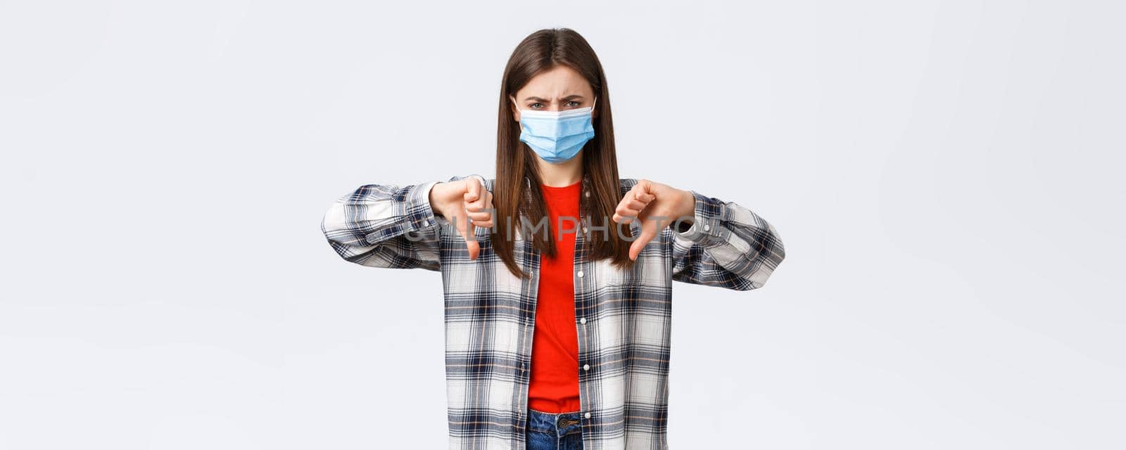 Coronavirus outbreak, leisure on quarantine, social distancing and emotions concept. Disappointed and mad young woman in medical mask vote against, show thumb-down and grimacing dismay.