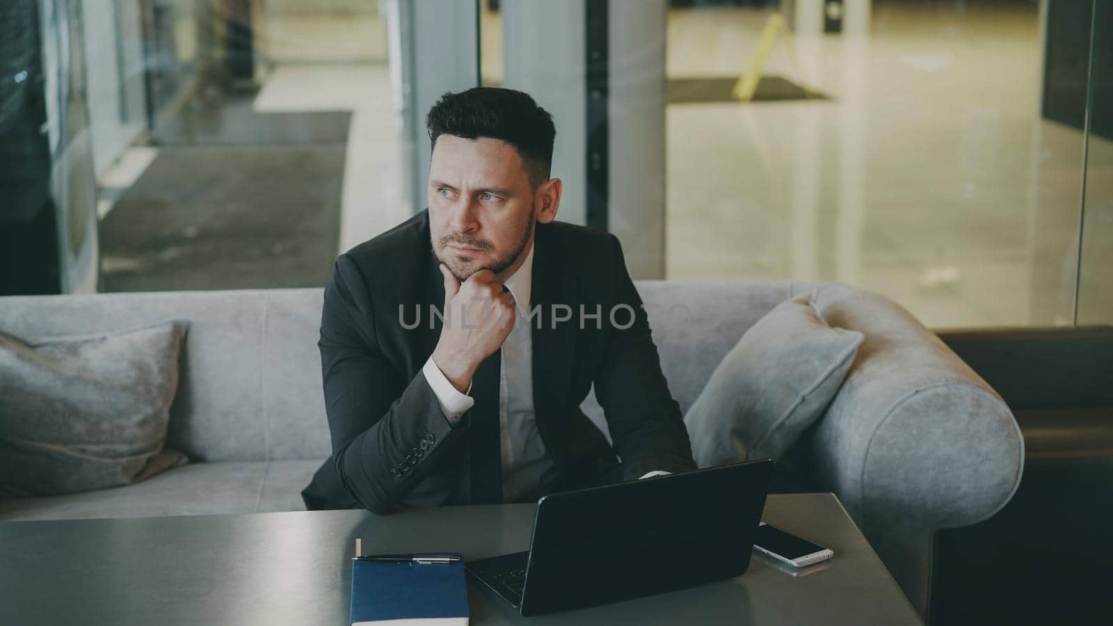 Upset thoughtful bearded Caucasian businessman sitting and thinking over his business plan while looking at his laptop screen in glassy cafe. Smartphone, notepad and coffee cup are on his table.