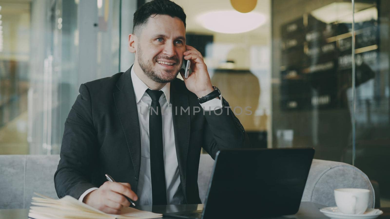 Portrait of cheerful Caucasian businessman sitting, smiling, talking on smartphone with his friend and writing down information in notepad while looking at laptop in modern cafe during lunch time