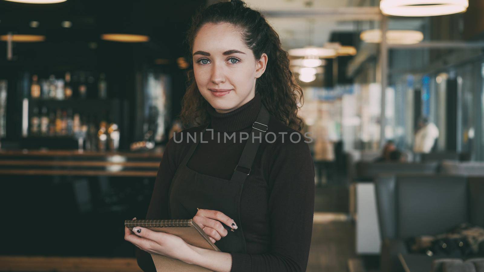 Portrait of cheerful and beautiful young waitress standing, keeping menu, and smiling sincerely in slylish cafe. Bar counter with many hard and soft drinks is in the background