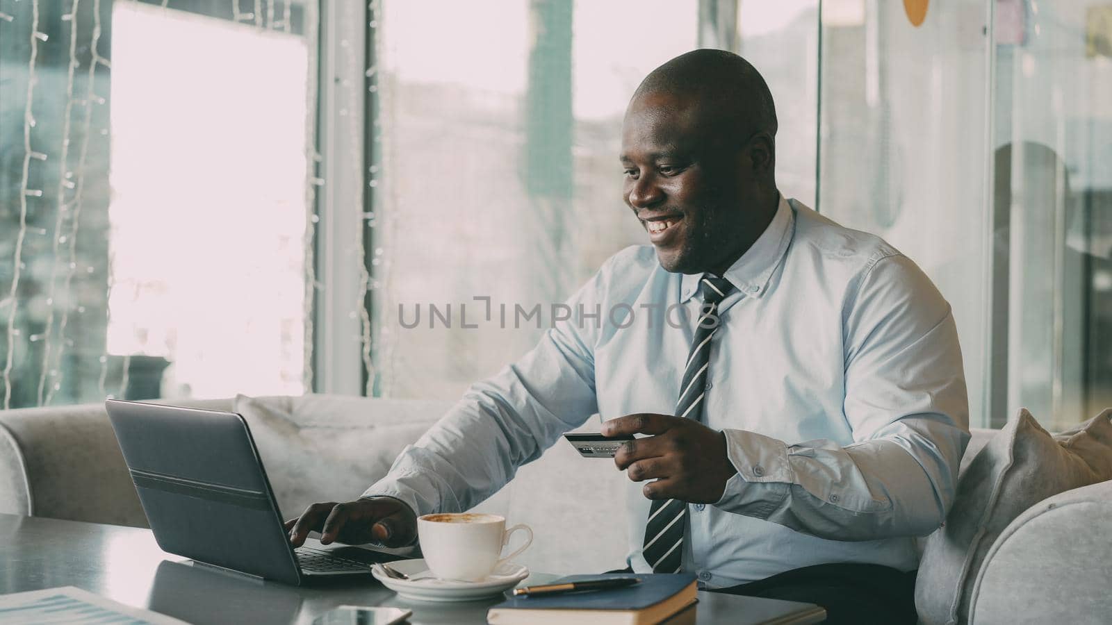Optimistic African American businessman in formal clothes paying online bill using his credit card and laptop in fashionable cafe during lunch breaak. He smiles happily and raises his clenched fist.