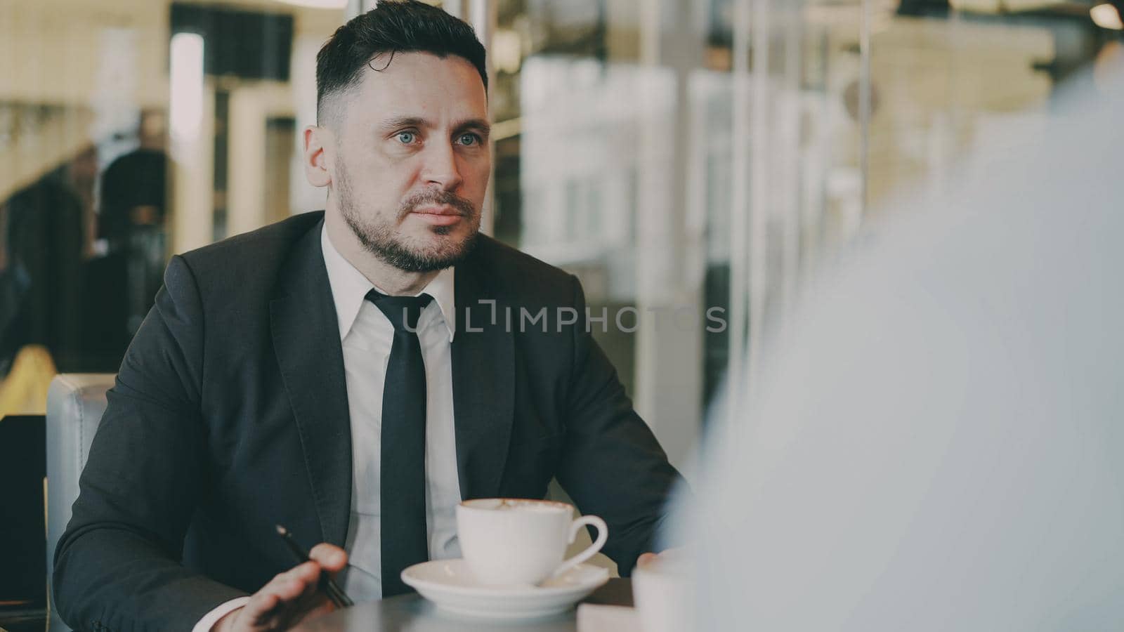 Bearded Caucasian entrepreneur gesticulating and discussing his startup plan with investor in formal wear in glassy cafe during during meeting