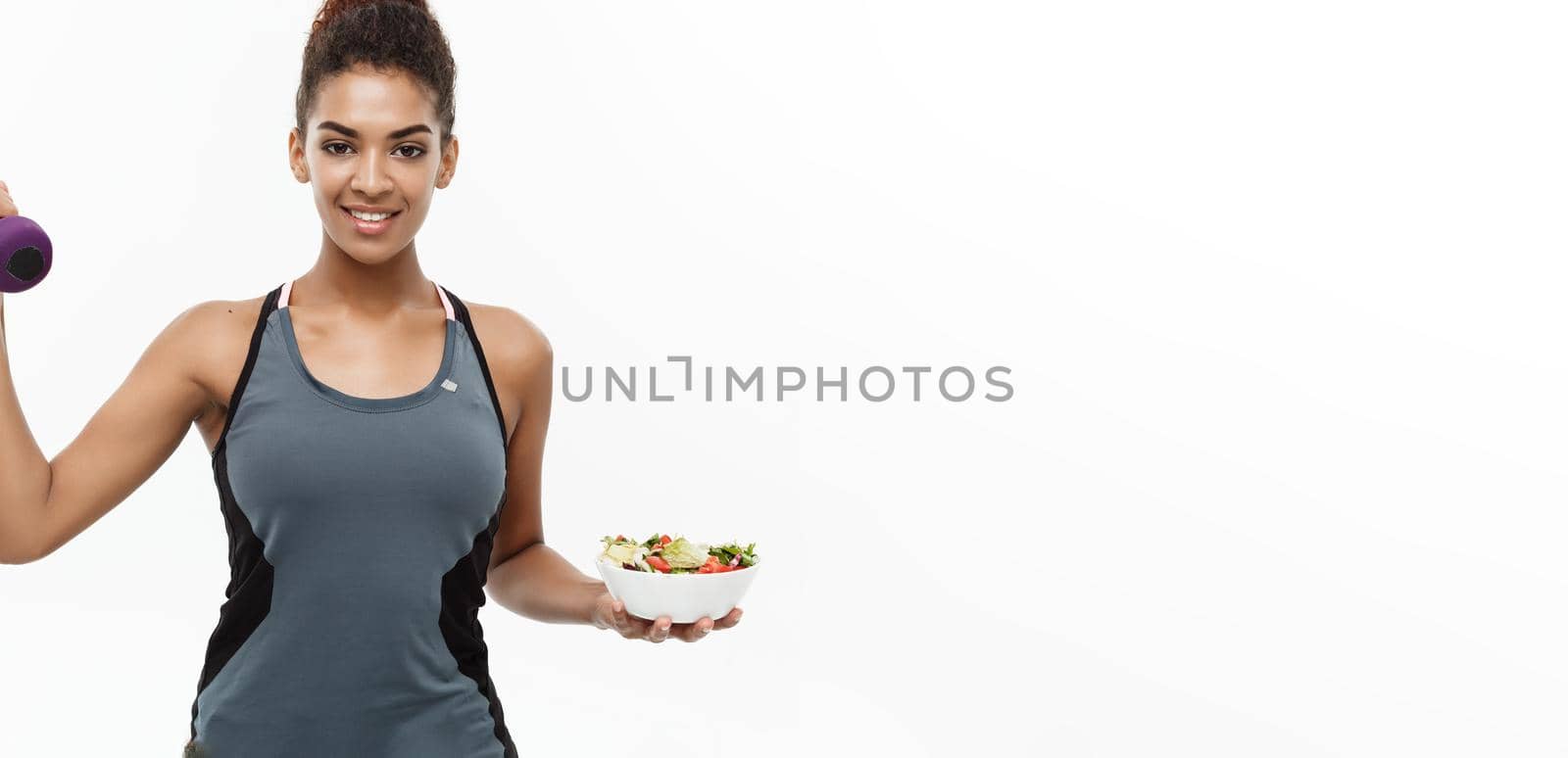 Healthy and Fitness concept - Beautiful sporty African American on diet holding dumbbell and fresh salad on hands. Isolated on white studio background