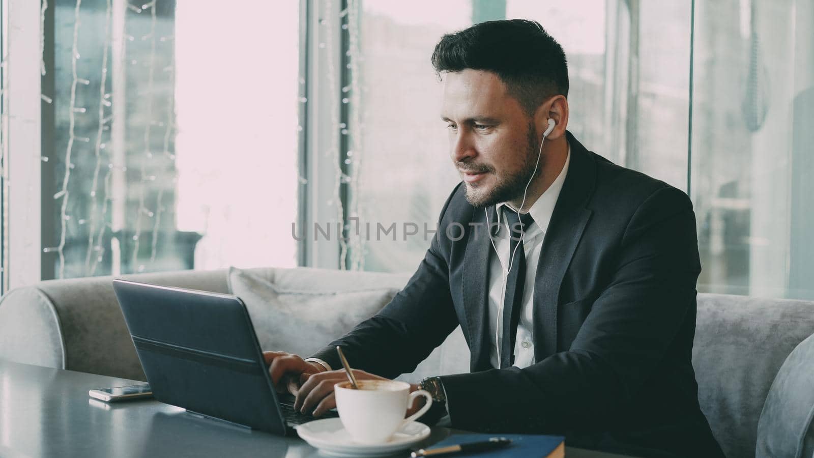 Happy Caucasian entrepreneur in formal clothes typing on his laptop while listening to music with earbuds in his ears in a modern cafe during lunch by silverkblack