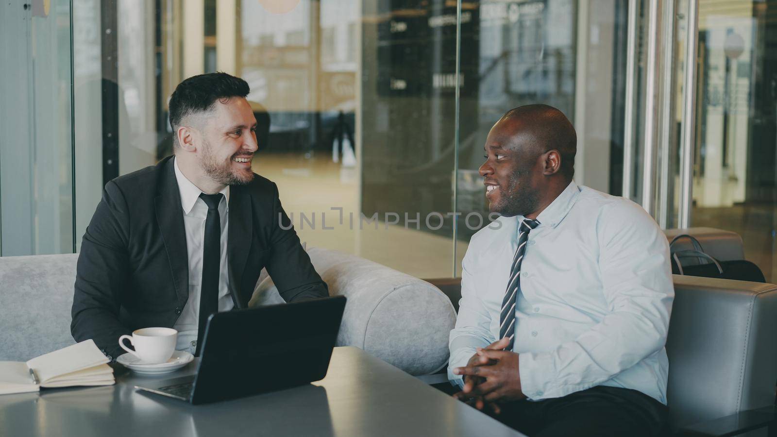 Two cheerful businessmen discussing project in modern glassy cafe. African American entrepreneur writing in notebook and his caucasian partner in business suit pointing at laptop computer