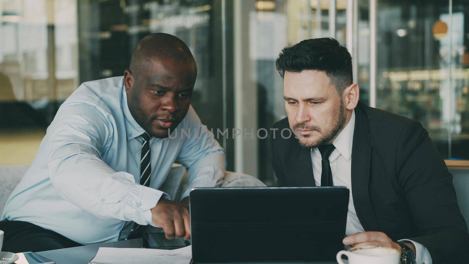 Two business colleagues looking at laptop computer and discussing their startup plan in modern office with glass walls. Bearded businessman and his partner sitting at table and talking future deals