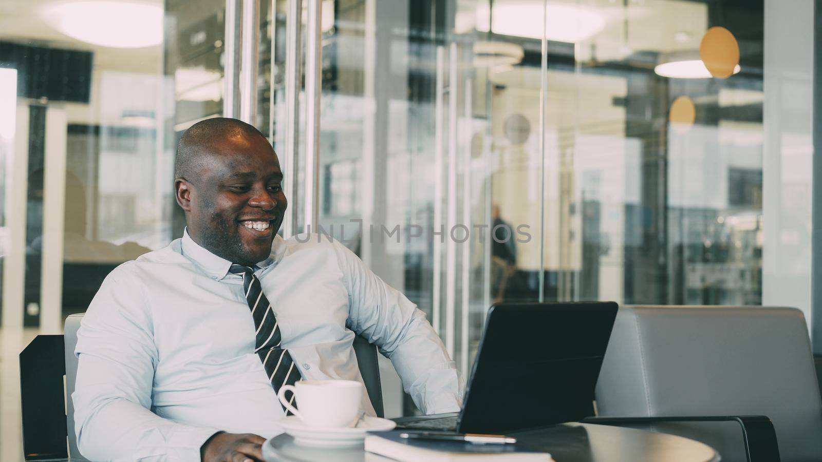 Portrait of hilarious African American businessman in formal clothes smiling, printing and working on his laptop in airy cafe during lunch break.
