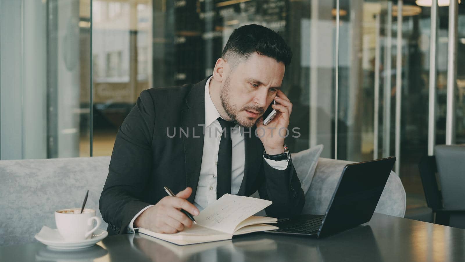 Bearded Caucasian businessman using laptop, talking on smartphone and writing down business information in modern cafe indoors during lunch break