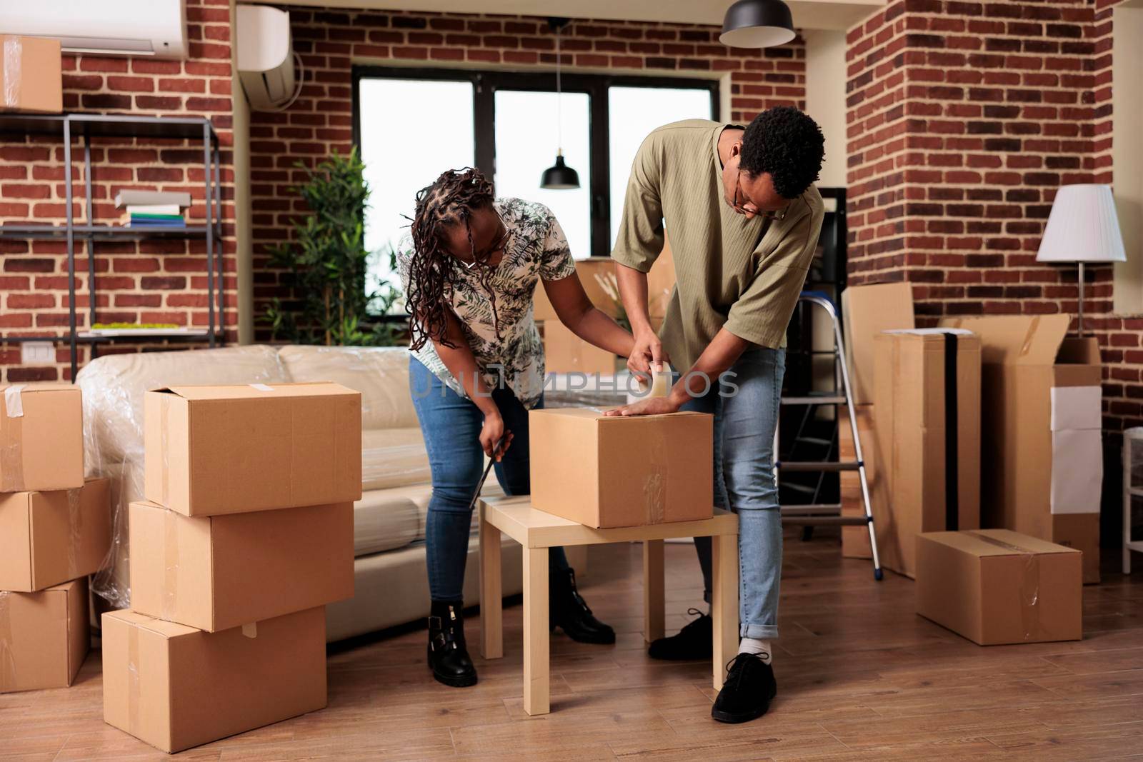 Married homeowners preparing furniture for transportation to new rented apartment flat, using sticky tape roller to pack things in boxes. Moving in family house bought on mortgage loan.