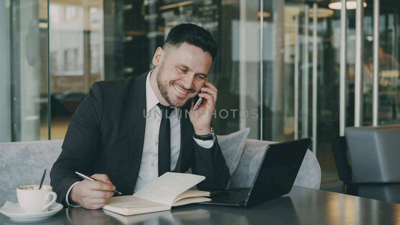 Bearded smiling businessman using laptop computer, talking on smartphone and writing down business information in modern cafe indoors during lunch break