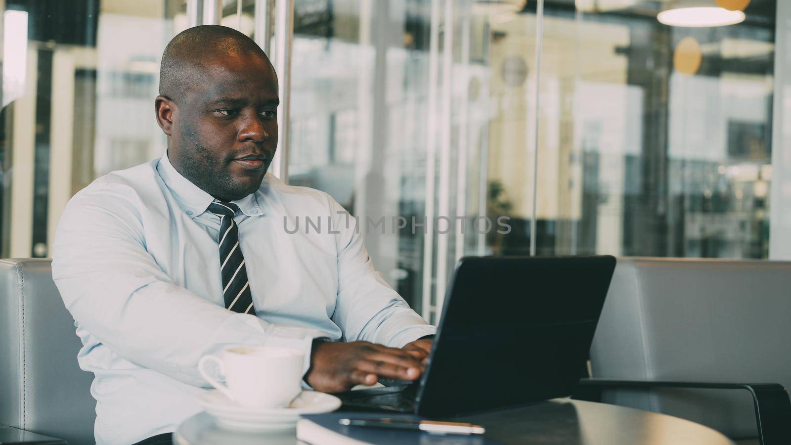 Portrait of African American businessman in formal clothes thinking, printing and working on his laptop in glassy cafe during lunch break. Coffee is on his table.