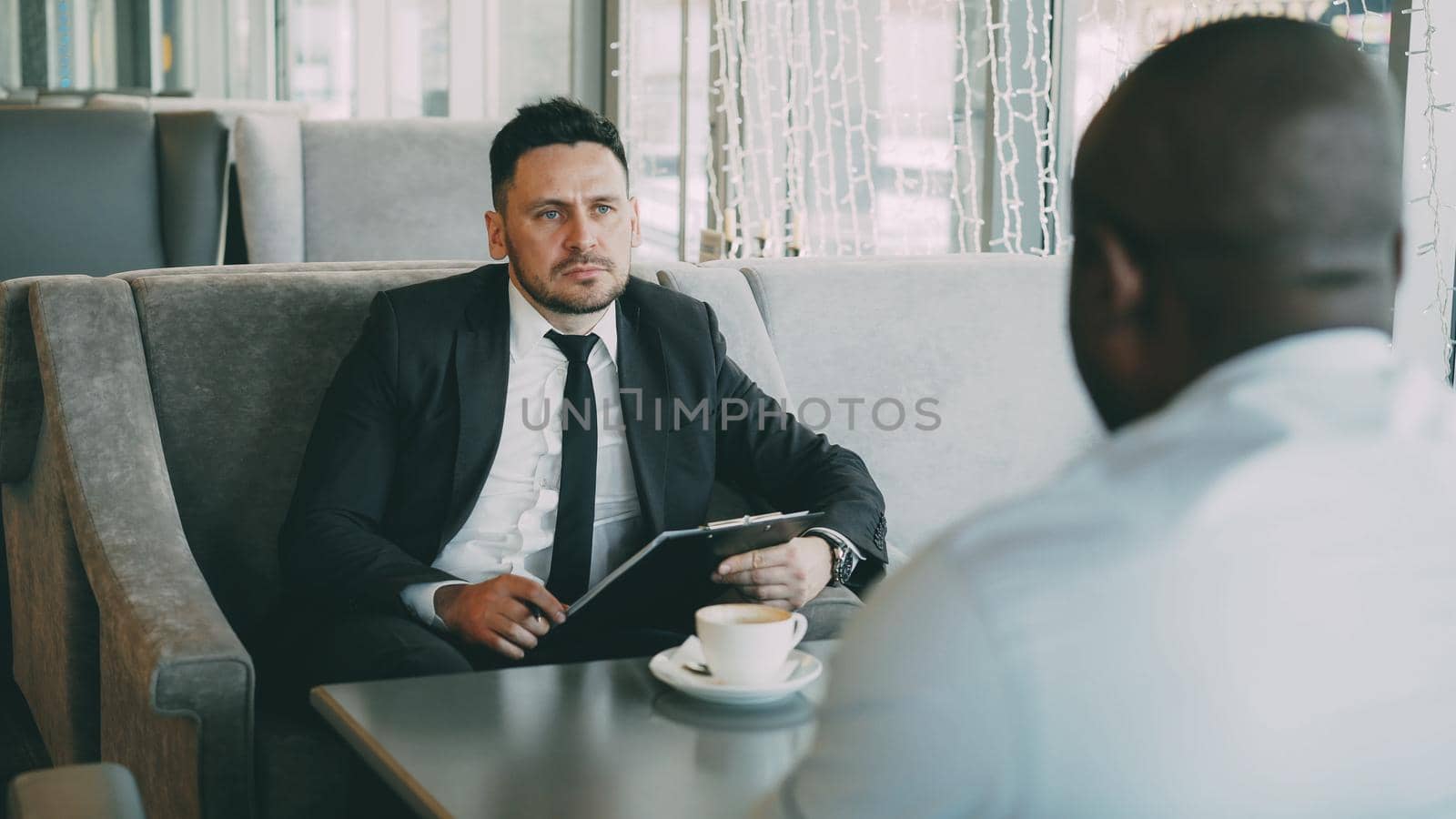 Portrait of bearded Caucasian businessman in formal clothes taking job interview with his Africcan American employee in glassy cafe during lunch break. Coffee cup and notepad are on his table
