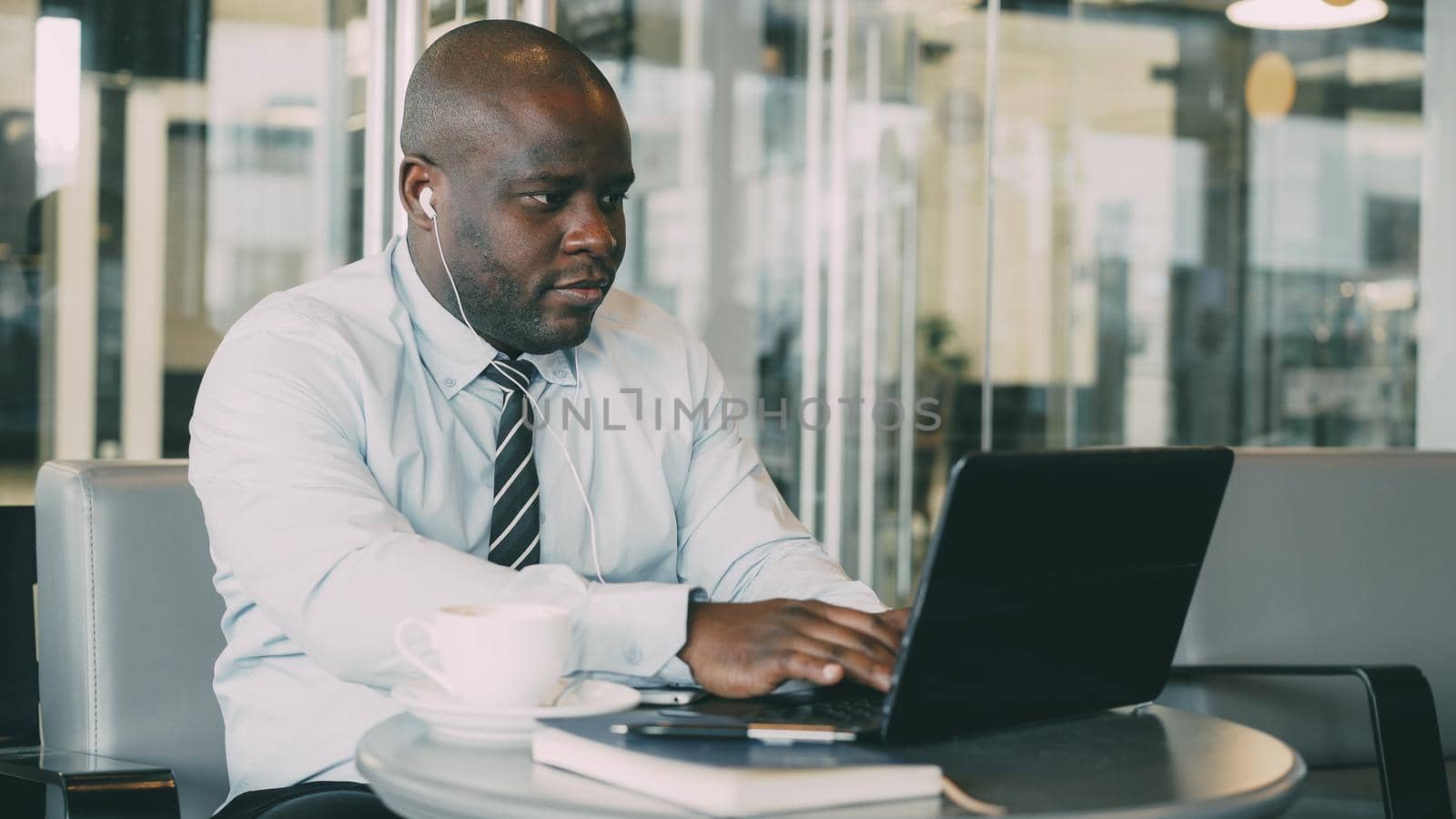 African American businessman in formal wear working and surfing social media on laptop while listening to music with earbuds in his ears in a glassy cafe by silverkblack