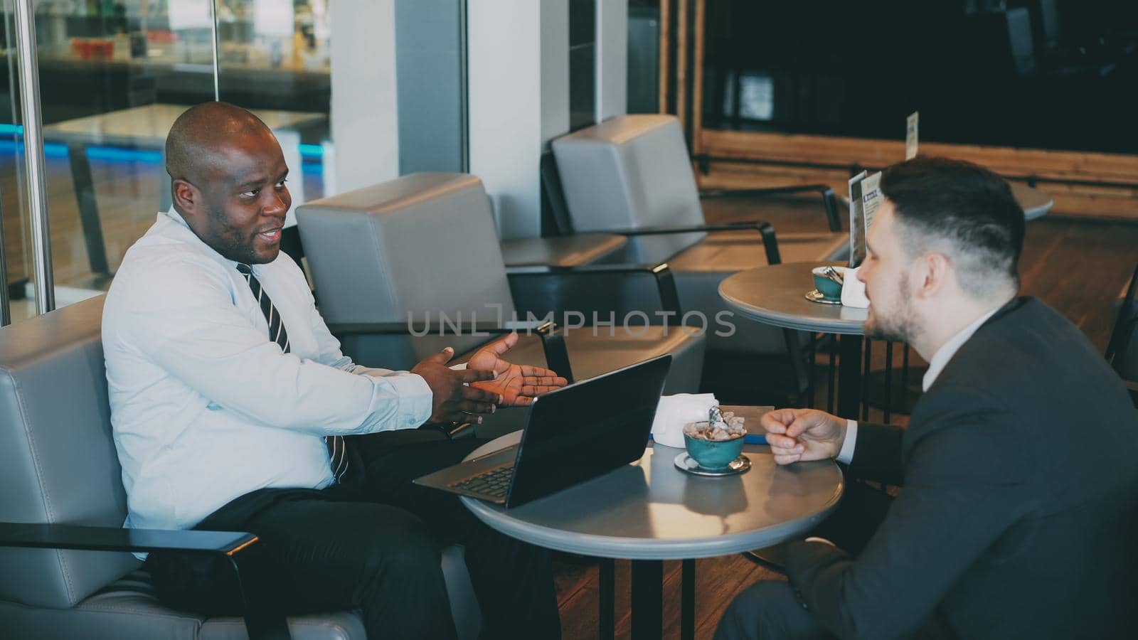 Two multiracial businessmen in formal clothes smiling, gesturing and discussing their startup in spacious cafe during lunch break. Advanced laptop is on their table. by silverkblack