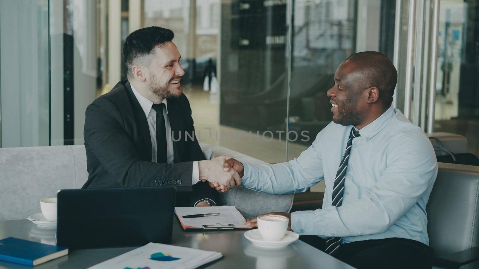 Two business colleagues discussing their business contract and looking at notepad in stylish cafe. African American businessman puts his signature and shakes the hand of Caucasian man by silverkblack
