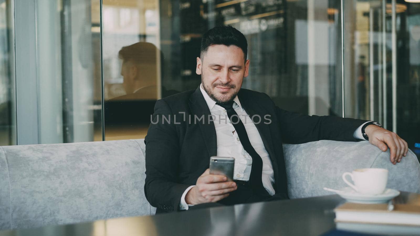 Smiling bearded Caucasian businessman in formal clothes smiling, relaxing and looking at his smartphone screen while drinking coffee in airy cafe during lunch time