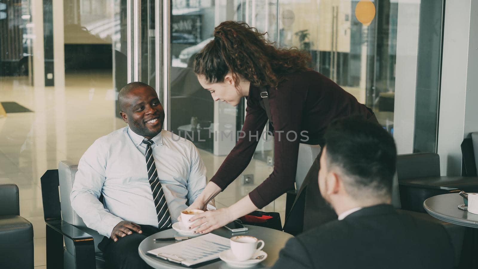Cheerful African American businessman in formal clothes smiling and discussing financial report with his Caucasian partner in cafe. Young waitress bringing cup of coffee. by silverkblack