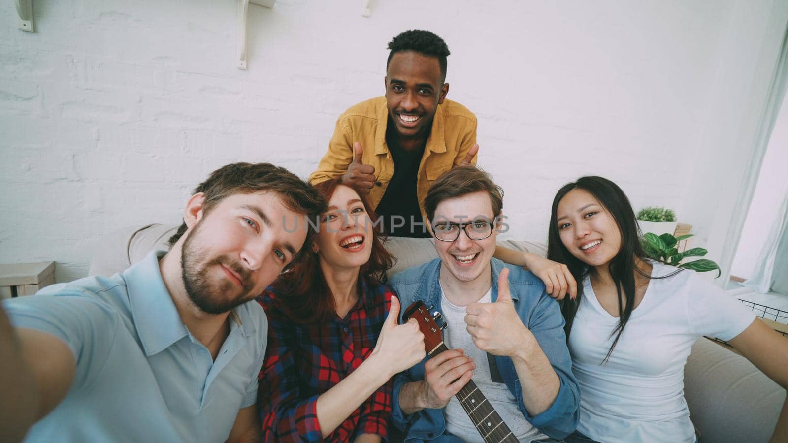 Point of view shot of multi ethnic group of cheerful friends taking selfie photos on smartphone camera while celebrating at party at home