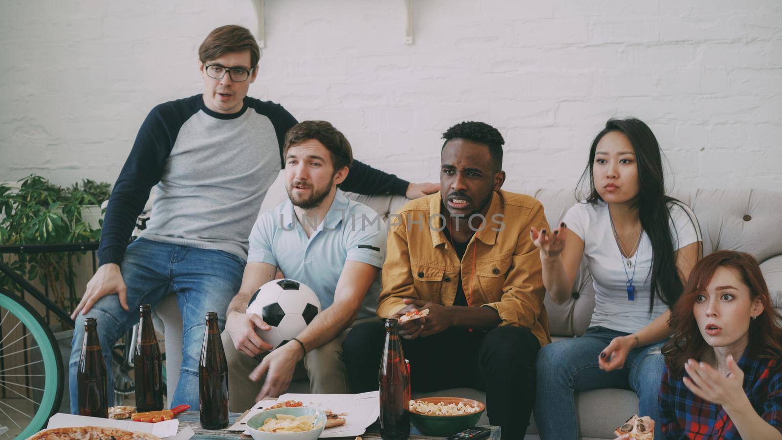 Multi ethnic group of friends sports fans watching football championship on TV together eating pizza and drinking beer at home indoors