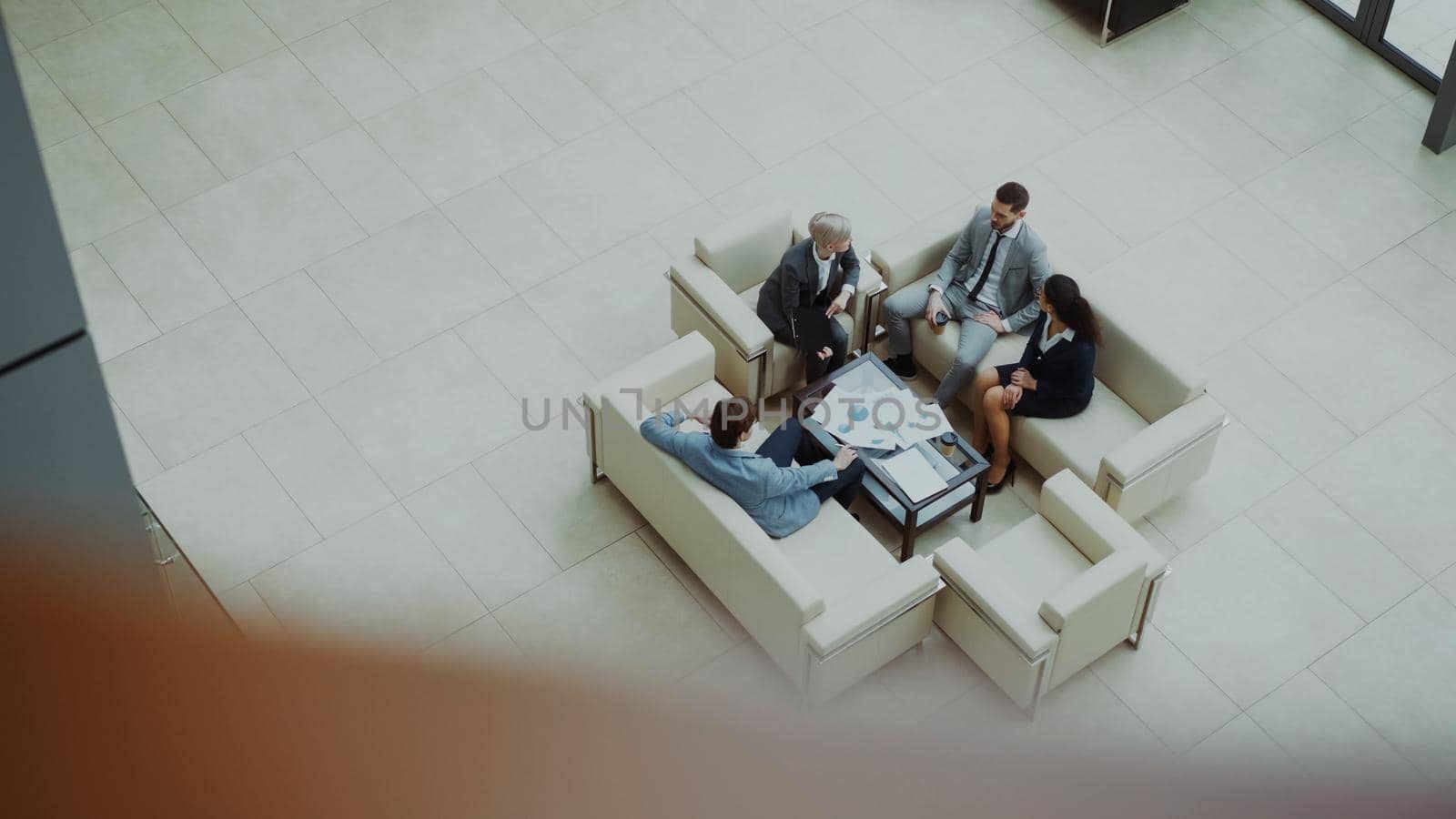 Top view of group of business people colleagues discussing financial charts sitting on couchs in lobby at modern business center by silverkblack