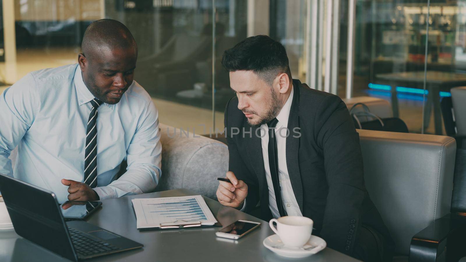 Two multi-ethnic partners in formal clothes sitting and discussing financial charts details while looking at notepad in glassy cafe during lunch time. by silverkblack
