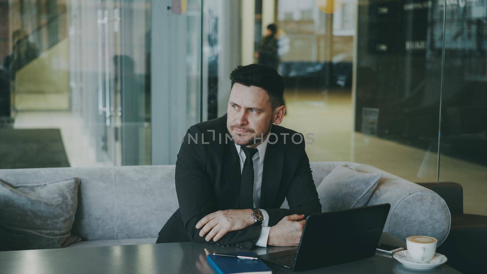 Thoughtful bearded Caucasian businessman sitting and thinking over his business plan while looking at his laptop screen in glassy cafe during lunch time by silverkblack