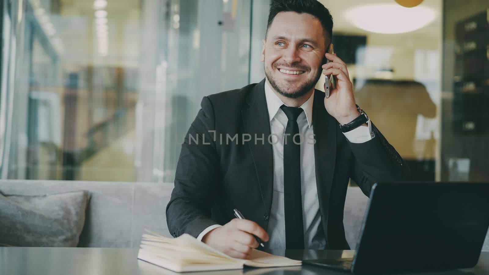 Happy bearded businessman in black business suit sitting at the table, smiling and talking on mobile phone in modern cafe. Man keeps a notebook at his hand