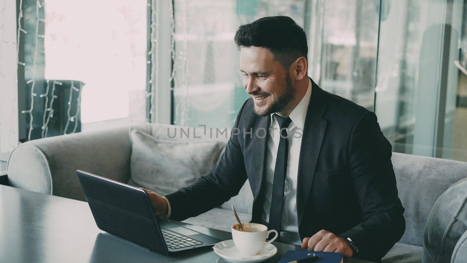 Caucasian businessman in formal clothes chatting through laptop webcam and waving his hand happily in a classy cafe during lunch time. by silverkblack