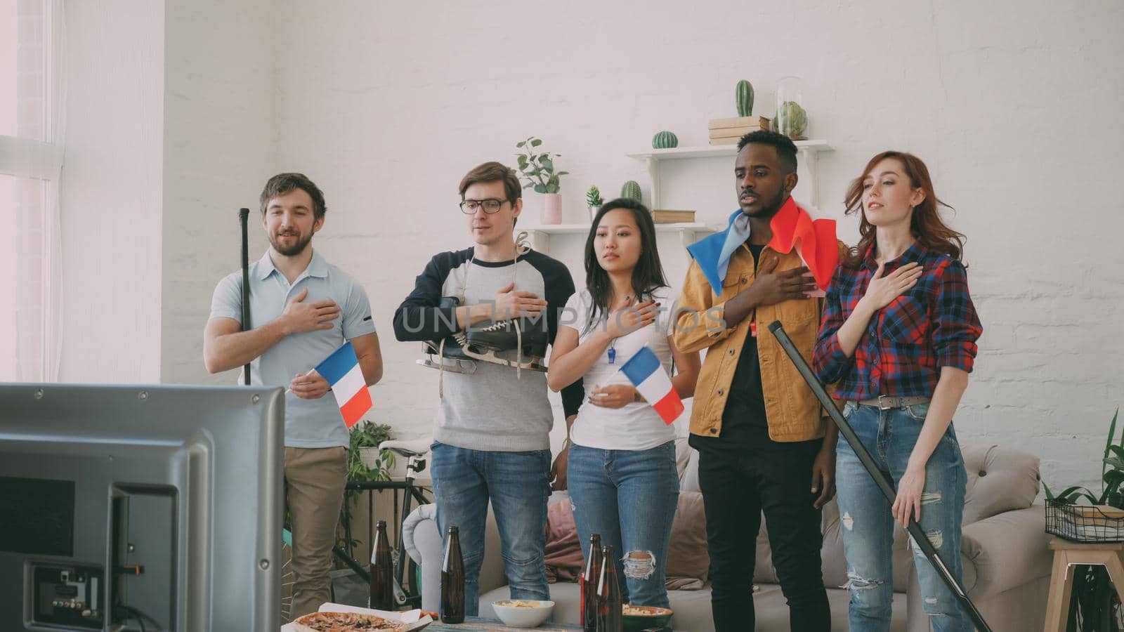 Multi ethnic group of friends sport fans listening and singing French national anthem before watching sports championship on TV together at home indoors