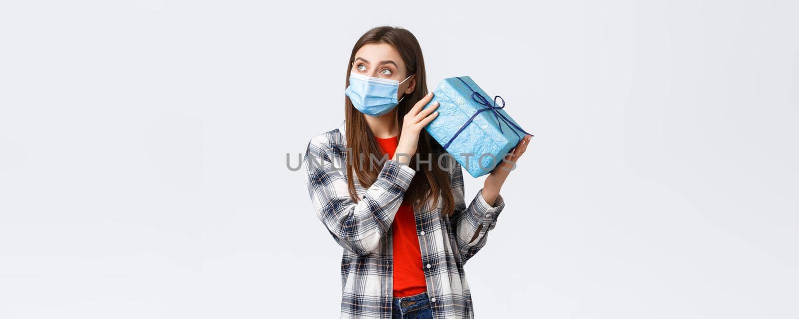 Covid-19, lifestyle, holidays and celebration concept. Curious young cute girl celebrating birthday, shaking gift box to guess what inside, look away thoughtful, lean ear to present, white background.