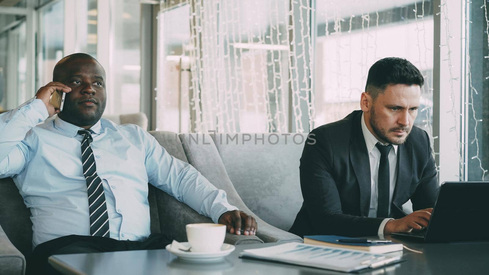 Bearded Caucasian businessman in formal clothes printing business information on his laptop and his African American partner talking on smartphone while sitting in stylish cafe during lunch.