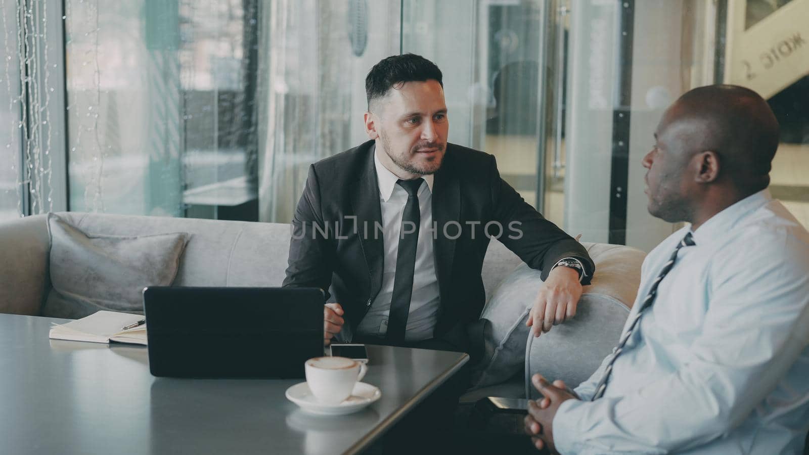 Two multiethnical partners in formal clothes looking at digital tablet and discussing their business ideas at a table with laptop and a cup of coffee in a trendy cafe by silverkblack