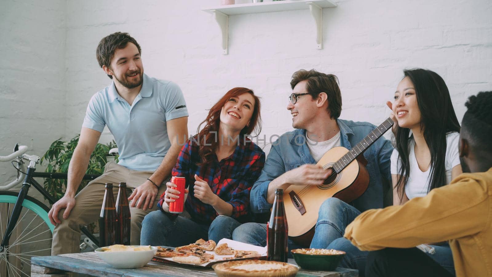 Happy young friends have party at shared apartment and singing together while their friend playing guitar at home indoors