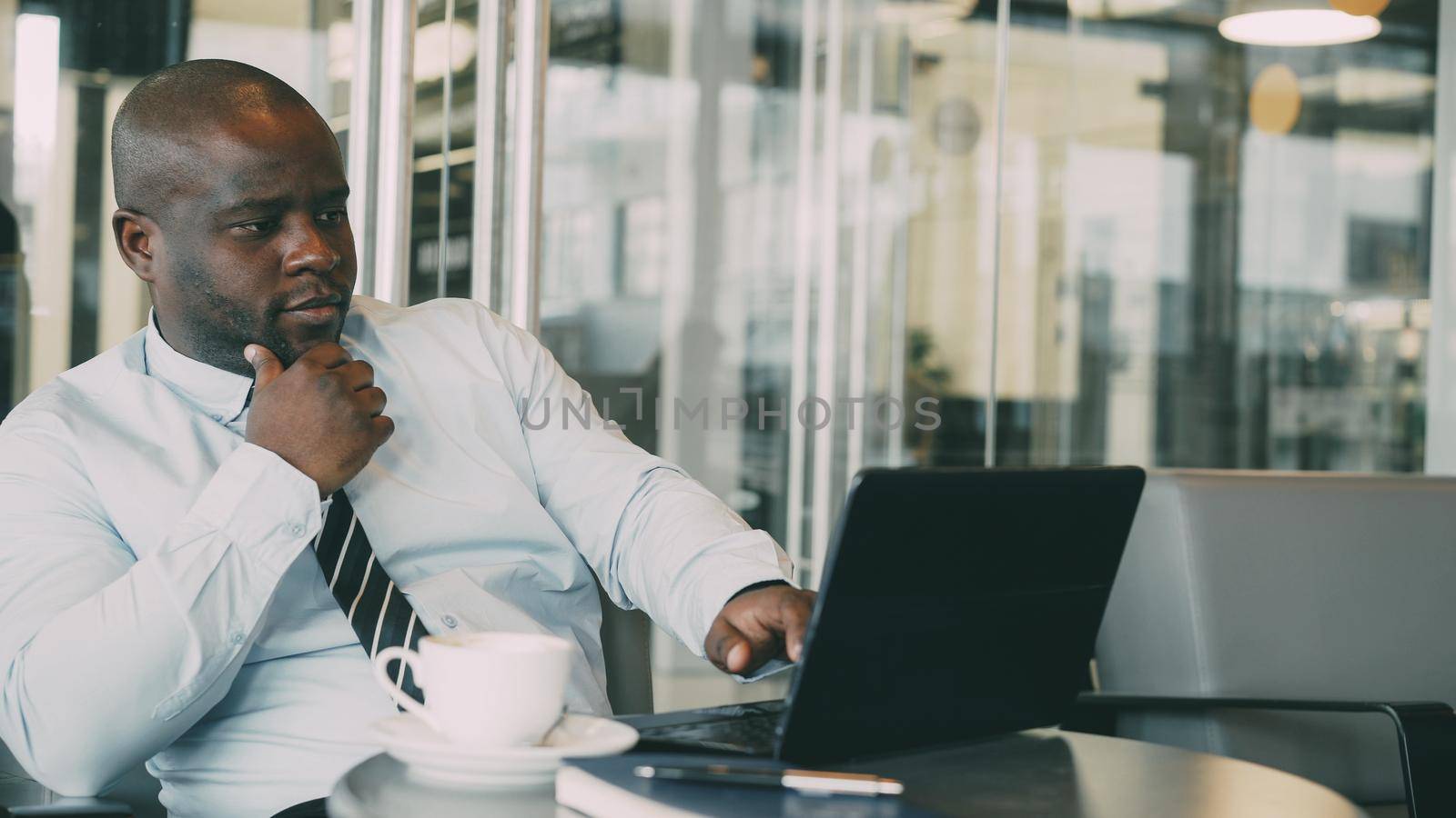 Portrait of African American businessman in formal clothes thinking, printing and working on his laptop in glassy cafe during lunch break. Coffee is on his table.