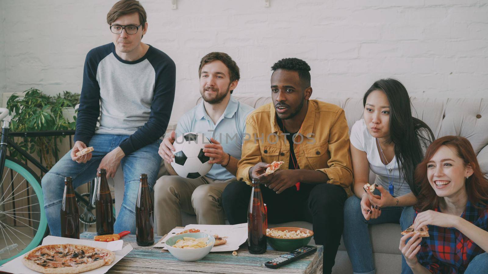 Multi ethnic group of friends sports fans watching football championship on TV together eating pizza and drinking beer at home indoors