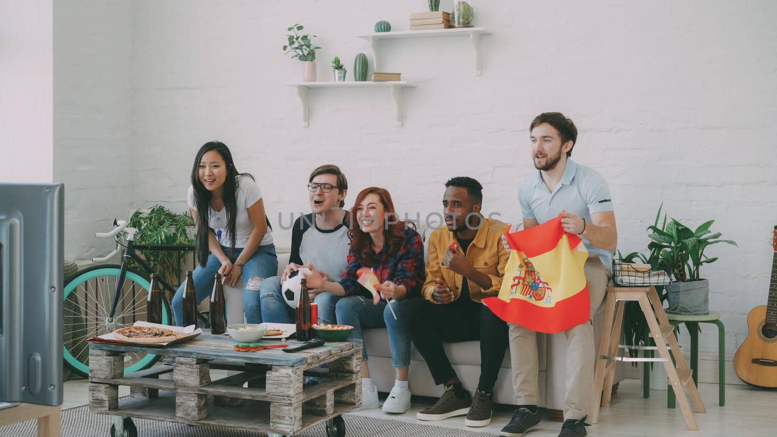 Multi-ethnic group of friends sports fans with Spanish flags watching football championship on TV together at home and cheering up favourite team by silverkblack