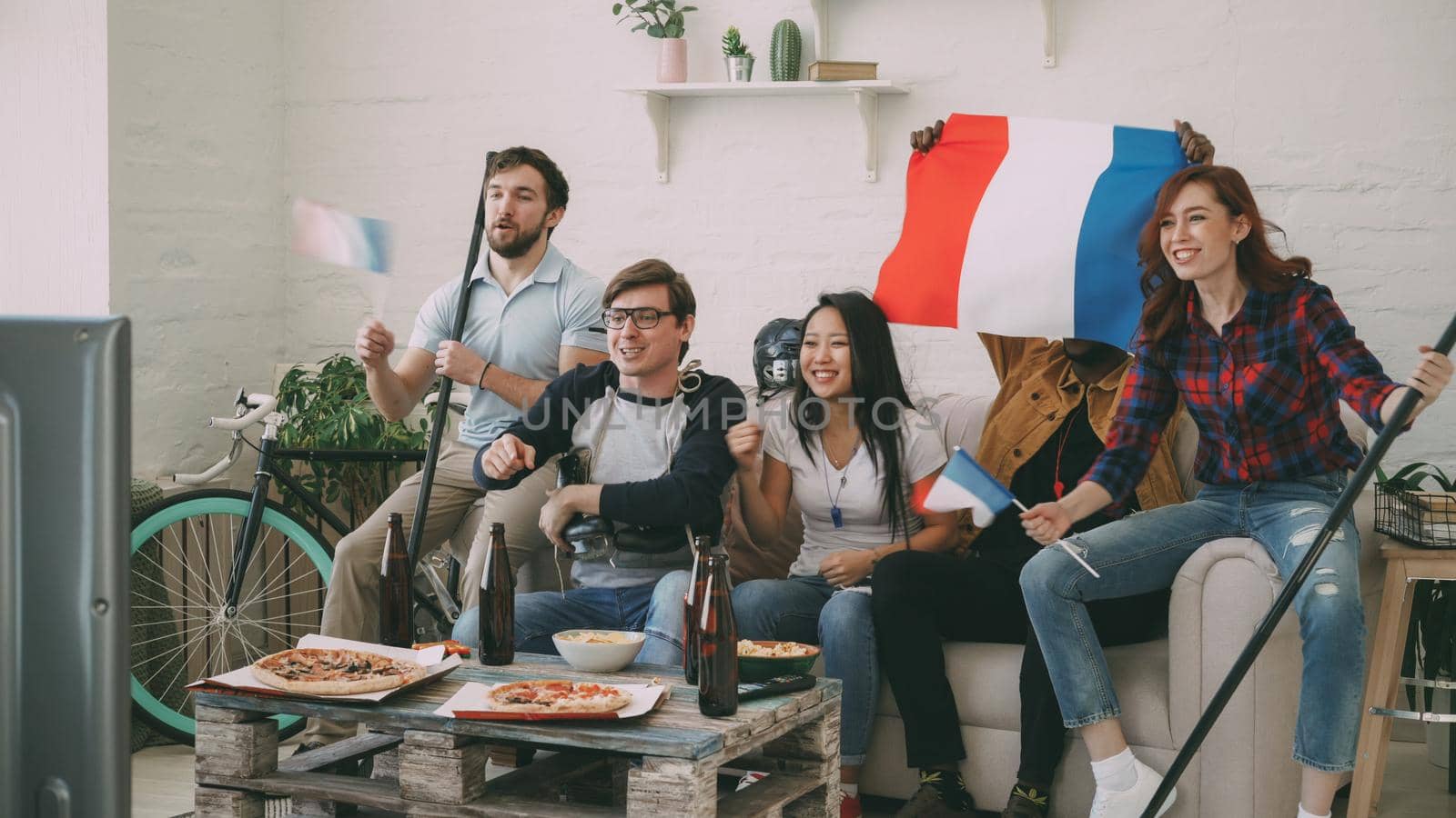 Group of young friends sports fans with French national flags watching sport championship on TV together cheering up favourite team at home indoors