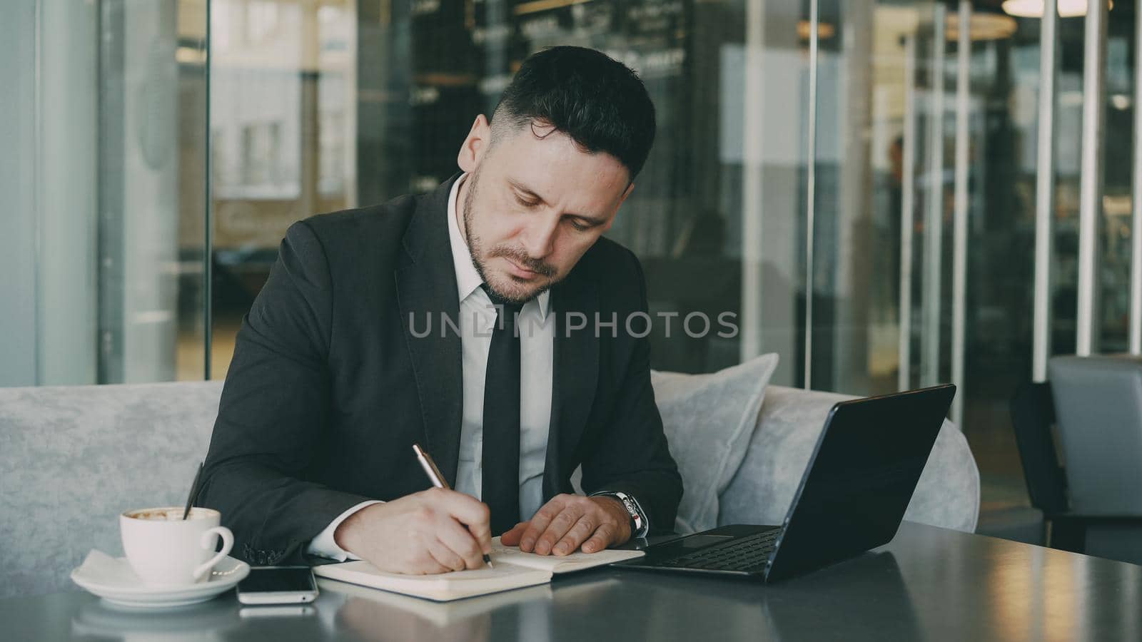 Smart Caucasian businessman using laptop computer and writing down information in his notepad in glassy cafe having coffee cup on his table. A cup of coffee is on his table.