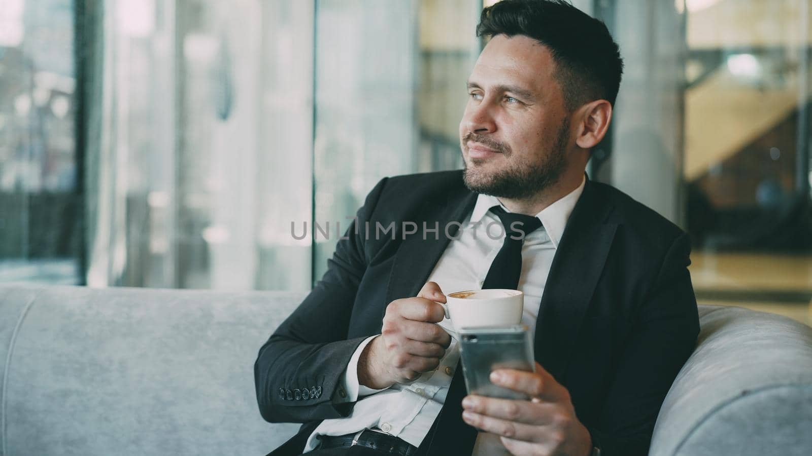 Happy Caucasian businessman in formal clothes smiling and looking at his smartphone screen while drinking coffee in airy cafe during lunch time by silverkblack