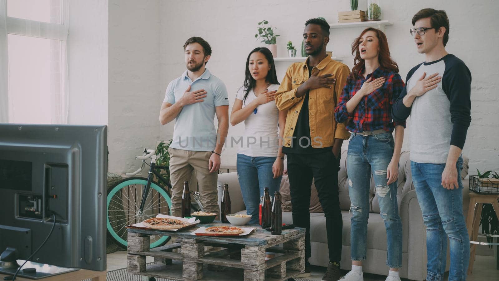 Multi ethnic group of friends sport fans singing national anthem before watching sports championship on TV together at home indoors