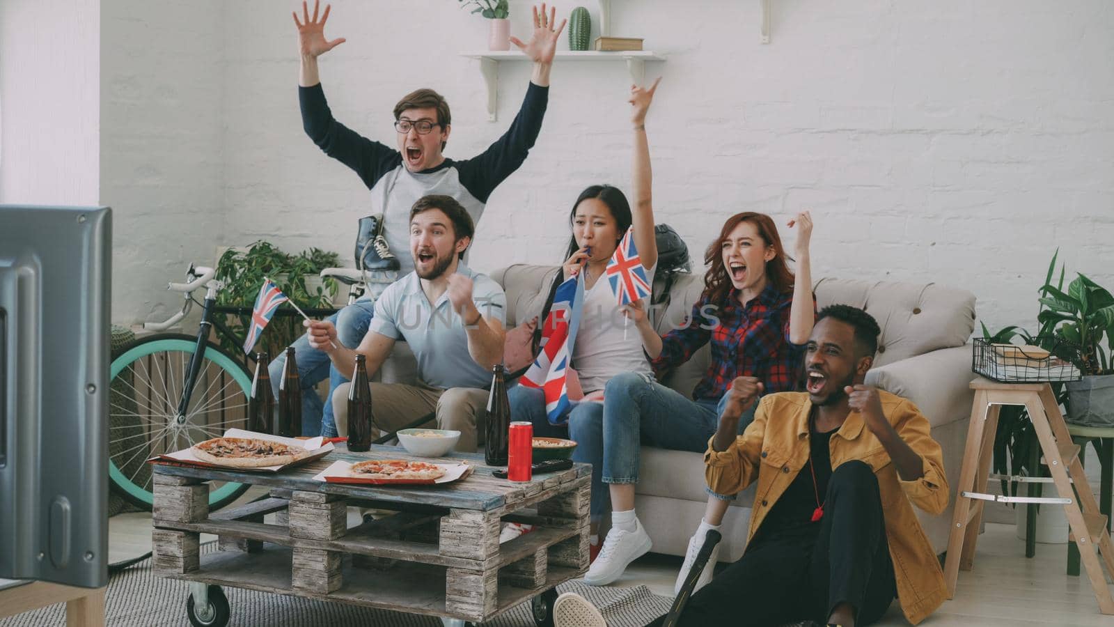 Multi ethnic group of friends sports fans with British national flags watching hockey championship on TV together cheering up their favourite team at home indoors