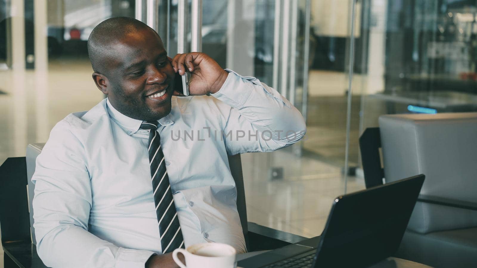 Portrait of African American businessman in formal clothes talking on his smartphone and browsing net while looking at his laptop in modern cafe during lunch time. He looks ambitious and successful.