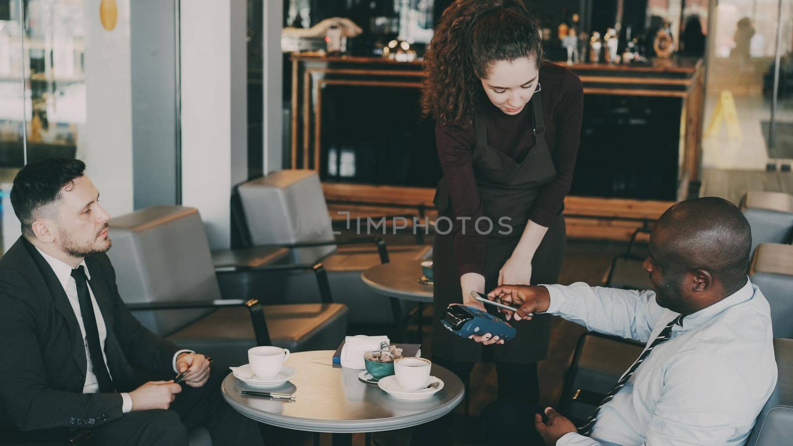 Two optimistic multiracial businessmen in formal wear pay online bill using smartphone in modern cafe during lunch break. Cheery young waitress standing and keeping portable cash terminal.